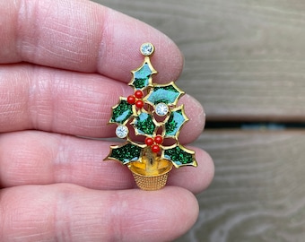 May include: A gold-coloured Christmas tree brooch with green glittered leaves, red berries, and clear crystal accents. The tree is in a woven basket. A festive holiday accessory.
