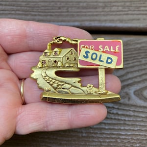 May include: A gold-toned brooch featuring a house with a winding road and a "FOR SALE SOLD" sign. The house has a chimney with smoke, and the sign is pink with blue lettering. The brooch is held in a hand.
