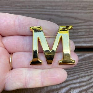 Może przedstawiać: Złota metalowa broszka w kształcie litery "M". Broszka ma szpilkę z tyłu do mocowania. Litera jest w kroju szeryfowym i ma błyszczące wykończenie.