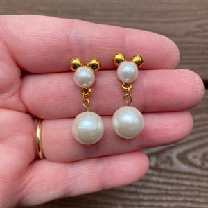May include: A pair of gold-toned earrings featuring a Mickey Mouse-inspired design. Each earring has a small gold top with three rounded shapes, connected by a gold chain to a large, round, white pearl. The earrings are held up by a person's hand.