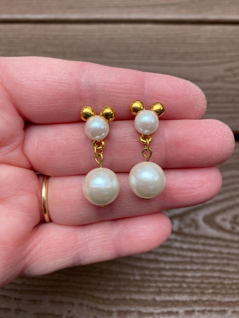 May include: A pair of gold-toned earrings featuring a Mickey Mouse-inspired design. Each earring has a small gold top with three rounded shapes, connected by a gold chain to a large, round, white pearl. The earrings are held up by a person's hand.
