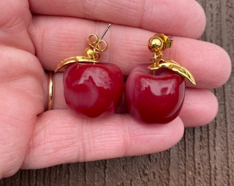 May include: A pair of red apple-shaped earrings with gold-colored leaf accents. The earrings have a post-back closure. The apples are a glossy, deep red color. The earrings are a fun and whimsical accessory.
