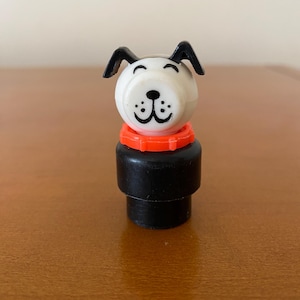 Puede incluir: Figura de perro de juguete vintage. El perro tiene una cabeza blanca con orejas negras y una nariz negra. Tiene un collar naranja y un cuerpo negro. El juguete es de plástico y es un juguete clásico.