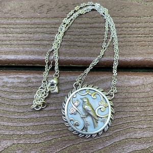 May include: A silver-toned necklace featuring a circular pendant with a mother-of-pearl inlay. The pendant displays a raised metallic bird perched on a branch, surrounded by decorative scrollwork and a twisted rope-style border on a thin chain.