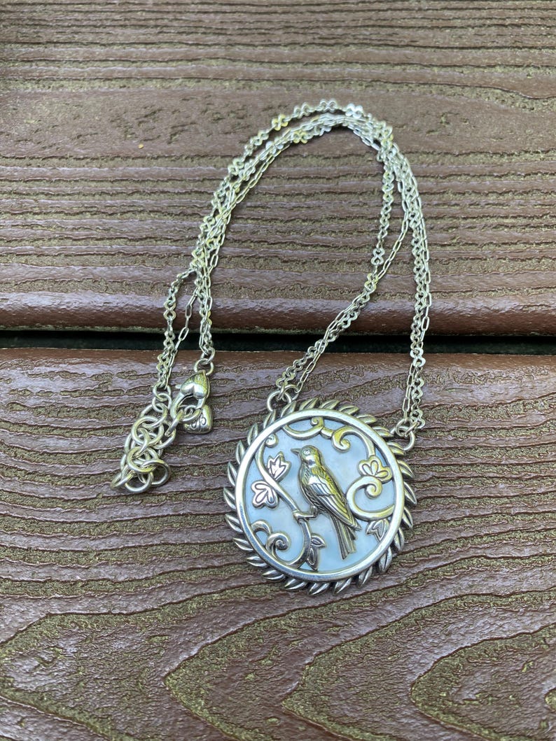 May include: A silver-toned necklace featuring a circular pendant with a mother-of-pearl inlay. The pendant displays a raised metallic bird perched on a branch, surrounded by decorative scrollwork and a twisted rope-style border on a thin chain.