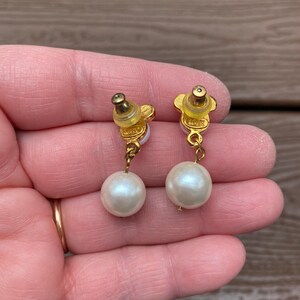 May include: A pair of gold-toned earrings featuring a flower-shaped stud and a dangling, large, round, white pearl. The earrings are held up by a person's hand.