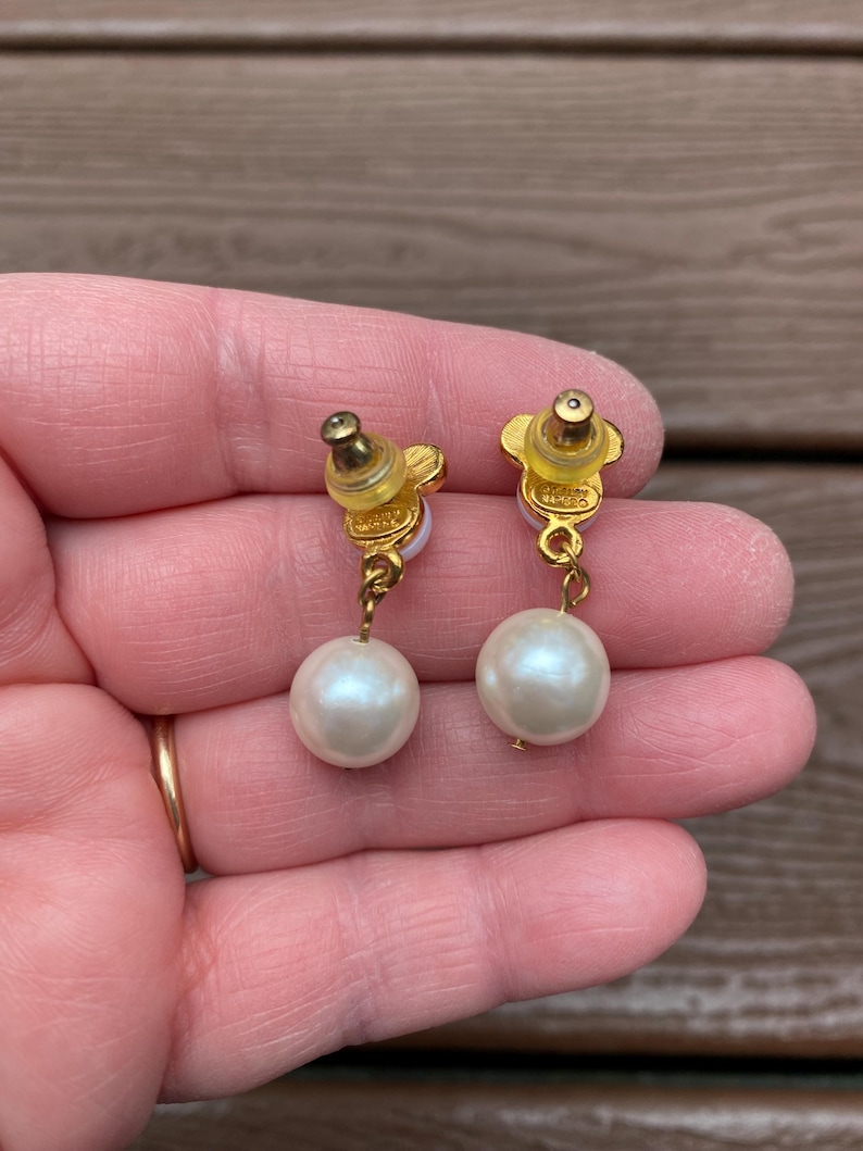 May include: A pair of gold-toned earrings featuring a flower-shaped stud and a dangling, large, round, white pearl. The earrings are held up by a person's hand.