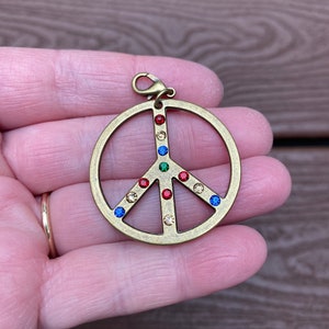 May include: A gold-colored peace sign pendant with a lobster clasp. The peace symbol is adorned with colorful gemstones in red, green, blue, and tan. The pendant is held in a hand.