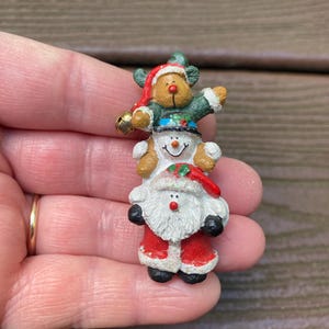 May include: A decorative brooch featuring a vertical stack of three festive figures. At the base is Santa Claus in a red suit, in the middle is a white snowman with a black hat, and at the top is a brown reindeer wearing a green jumper and red hat.