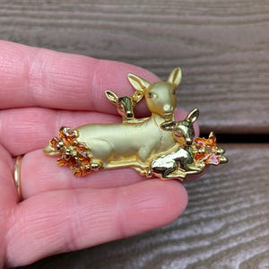 May include: A gold-coloured brooch depicting a mother deer and two fawns. The brooch has a matte finish and is decorated with orange and gold floral elements. The design is a charming representation of wildlife.