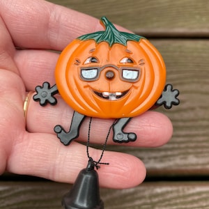 May include: A vintage Halloween pumpkin pin with a smiling face, glasses, and black arms and legs. The orange pumpkin has a green stem and a black bell attached by a string. A fun, festive accessory.