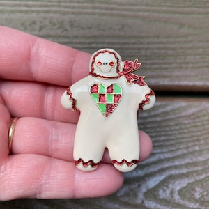 May include: A cream-colored gingerbread person brooch featuring red glitter accents on the edges, a red glitter bow at the neck, and a green and red checkered heart on the chest. The figure has two small red dots for eyes and a simple smiling mouth.