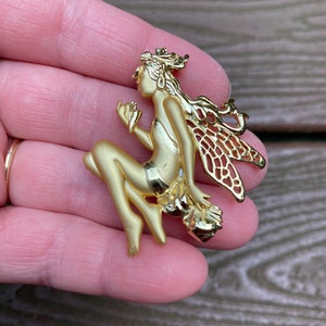 Pode incluir: Uma figura de fada dourada com asas, uma coroa e uma flor na mão. A fada está sentada em uma rocha.