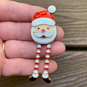 May include: A Santa Claus brooch with a red hat and white beard. The brooch has gold trim and red and white striped legs with black shoes. The Santa face is white with a rosy nose and a gold moustache.
