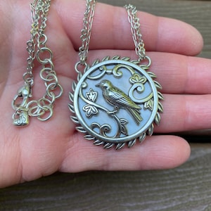 May include: A silver-toned necklace featuring a circular pendant with a mother-of-pearl inlay. The pendant depicts a bird perched on a branch, surrounded by a decorative leaf-patterned border. The chain includes a lobster clasp and a heart-shaped charm.