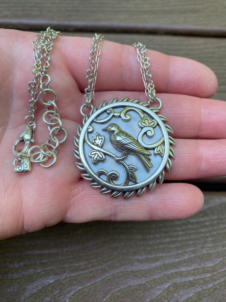 May include: A silver-toned necklace featuring a circular pendant with a mother-of-pearl inlay. The pendant depicts a bird perched on a branch, surrounded by a decorative leaf-patterned border. The chain includes a lobster clasp and a heart-shaped charm.