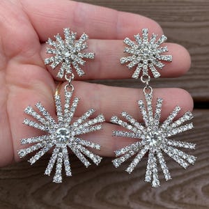 May include: A pair of silver-toned, starburst-shaped earrings. Each earring features a cluster of small, clear crystals at the center, with radiating crystal-studded arms. The earrings are designed to dangle, adding a touch of sparkle.