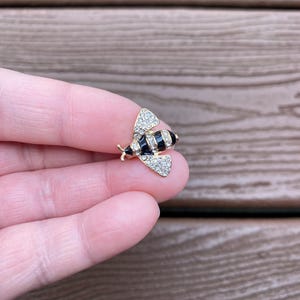 May include: A gold-toned bee brooch with black and white striped body and crystal-studded wings. The brooch is small and detailed, with a classic design. The bee's wings are adorned with small, sparkling crystals, adding a touch of elegance.