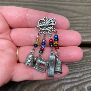 May include: A silver-toned sewing-themed charm brooch featuring a top piece shaped like scissors. Four dangling chains are attached, each adorned with colourful glass beads and a miniature metal charm: a thimble, a sewing machine, a ruler, and a spool.