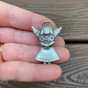 Pode incluir: Um pequeno pingente prateado em forma de anjo. O anjo tem asas detalhadas, uma aur&eacute;ola e segura um buqu&ecirc; de flores. O pingente &eacute; uma figura em miniatura, provavelmente para joias ou artesanato.