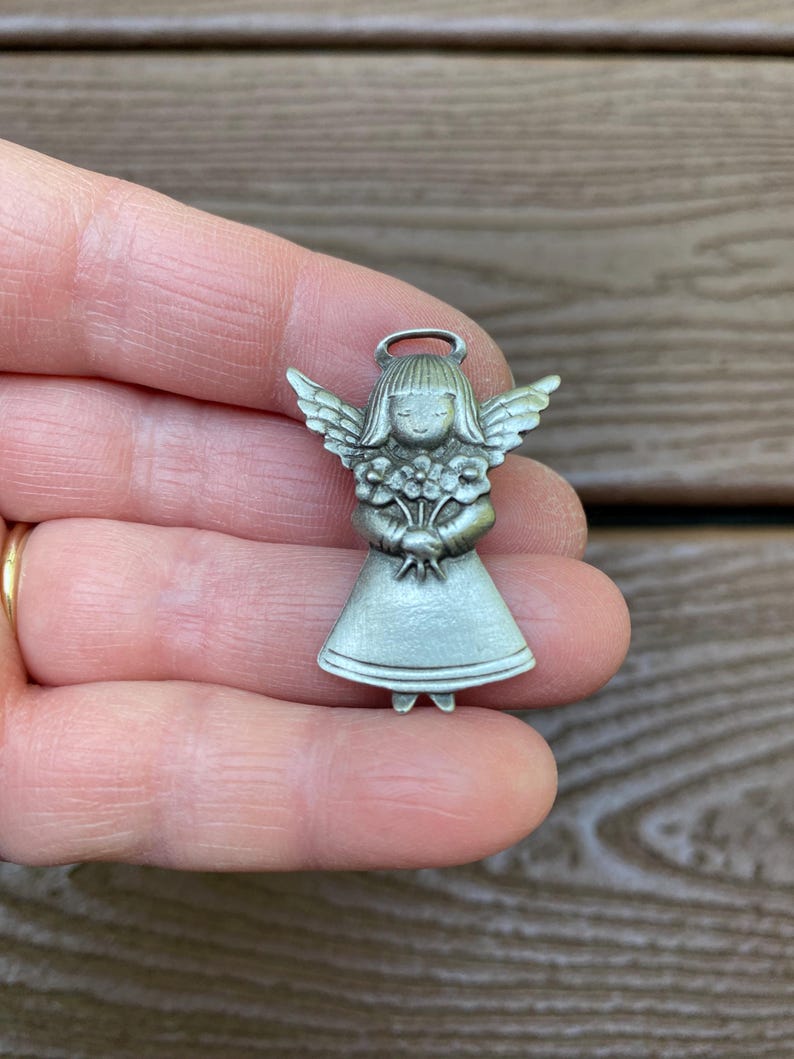 Pode incluir: Um pequeno pingente prateado em forma de anjo. O anjo tem asas detalhadas, uma aur&eacute;ola e segura um buqu&ecirc; de flores. O pingente &eacute; uma figura em miniatura, provavelmente para joias ou artesanato.