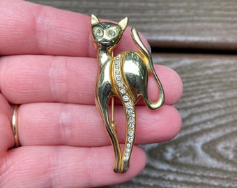 May include: A gold-toned cat brooch featuring a sleek, standing feline silhouette. The cat's eyes are accented with small clear crystals, and a vertical row of sparkling clear rhinestones runs down the centre of its body, highlighting the form.