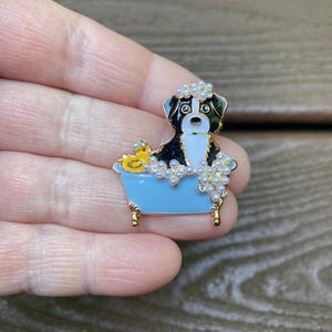 May include: A gold-tone metal brooch featuring a black and white dog sitting in a light blue bathtub. The dog is surrounded by white pearl-like bubbles and a small yellow rubber duck is positioned on the edge of the tub with gold-tone feet.
