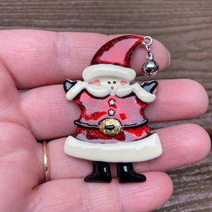May include: A Santa Claus brooch featuring a red glittery hat and coat, a cream-coloured beard and trim, a gold glittery belt buckle, and black boots. A small silver-toned metal jingle bell hangs from the tip of the Santa hat by a jump ring.
