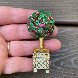 Könnte beinhalten: Eine goldfarbene Brosche in Form eines Formgehölzes in einem rechteckigen Pflanzgefäß. Der Baum hat grün emaillierte Blätter und runde rosa Strasssteine. Das Pflanzgefäß ist cremefarben mit goldenem Gittermuster und zwei kleinen bernsteinfarbenen Steinen.