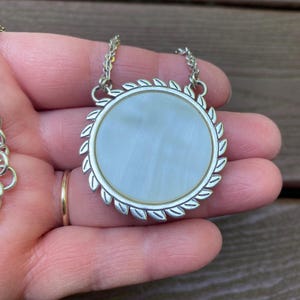 May include: A round pendant necklace featuring a smooth, iridescent white mother-of-pearl inlay set within a silver-toned metal frame. The circular border is decorated with a repeating leaf-like pattern, and the pendant hangs from a silver chain.