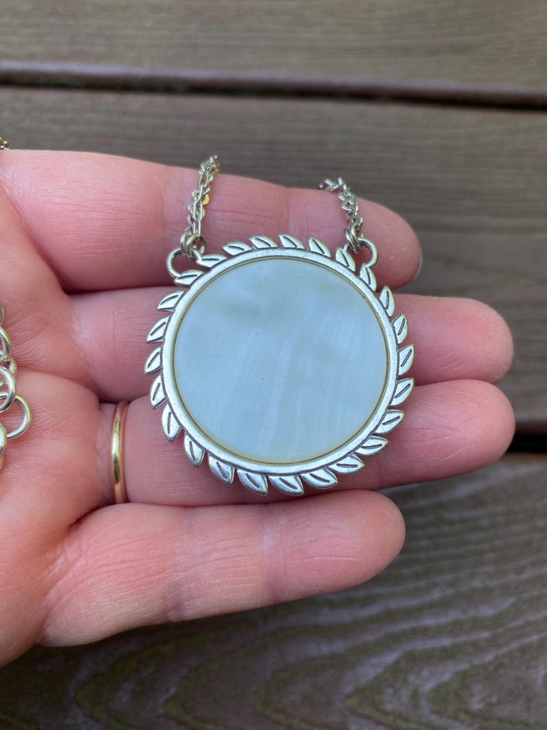 May include: A round pendant necklace featuring a smooth, iridescent white mother-of-pearl inlay set within a silver-toned metal frame. The circular border is decorated with a repeating leaf-like pattern, and the pendant hangs from a silver chain.