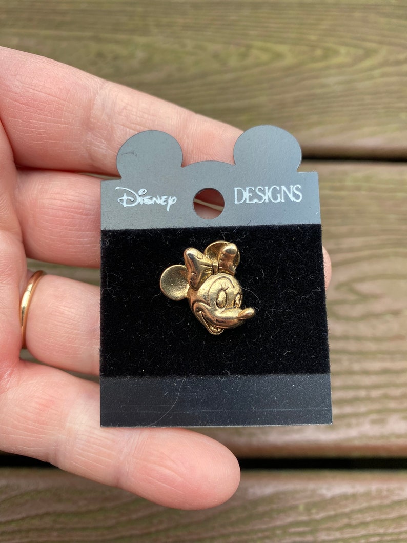 May include: A gold-coloured Minnie Mouse pin on a black velvet backing, attached to a grey Disney Designs card. The pin features Minnie's iconic bow and smiling face. The card has a Mickey Mouse head shape at the top.