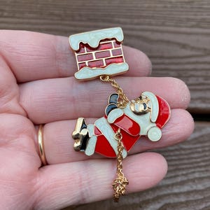 Puede incluir: Un broche navideño con Papá Noel bajando por una chimenea. El broche tiene una combinación de colores rojo, blanco y negro con detalles dorados. La chimenea es de ladrillo rojo con nieve blanca. Papá Noel lleva un traje y un gorro rojos.