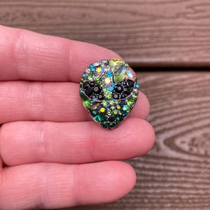 以下が含まれることがあります： さまざまな色のラインストーンが特徴の、キラキラ光るエイリアンの頭のピン。エイリアンの目は黒で、頭は緑、青、透明のクリスタルで飾られています。木製の背景を背景に、手でピンを持っています。