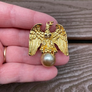 May include: A gold-toned eagle brooch with outstretched wings and a pearl-like orb. The eagle has detailed feathering and a small red accent on its head. The brooch is held in a hand, showcasing its intricate design.