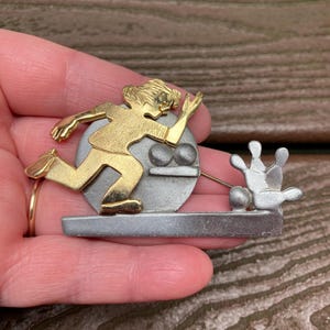 May include: A gold-toned metal brooch depicting a stylised figure in a running or bowling motion, positioned in front of a silver-toned circular disc. The design includes a silver-toned bowling lane base with a bowling ball and pins.