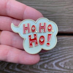 May include: A cloud-shaped enamel pin with a cream-coloured background and gold-toned metal edges. The text "Ho, Ho, Ho!" is written in bold, red, capital letters across the centre of the pin, mimicking a comic book speech bubble design.