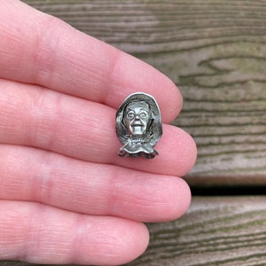 May include: A silver-toned metal pin featuring a detailed depiction of a character from the movie "The Wizard of Oz". The character is wearing a hat and has a surprised expression on their face.