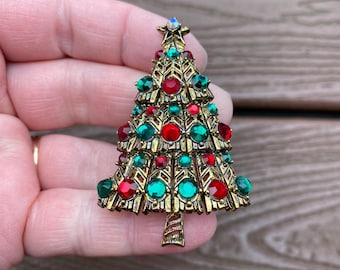 May include: A gold-toned Christmas tree brooch adorned with red and green gemstones. The brooch features a star at the top and a textured trunk. A festive accessory for the holiday season.