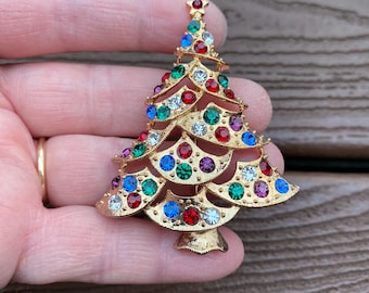 Può includere: Spilla a forma di albero di Natale color oro ornata di strass colorati. La spilla presenta una stella in cima ed è impreziosita da gemme rosse, verdi, blu e trasparenti. Un accessorio festivo.