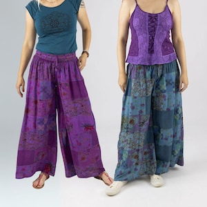 Op de afbeelding: Twee vrouwen in kleurrijke patchworkbroeken en tops. De ene vrouw draagt een teal top met een mandala-ontwerp en een paarse broek. De andere draagt een paarse top met vetersluiting en een blauwe patchworkbroek. De broeken zijn wijd.