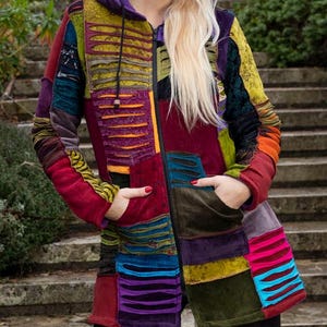 Może przedstawiać: Wielokolorowy płaszcz patchworkowy z kapturem, w odcieniach czerwieni, zieleni i fioletu. Płaszcz ma zamek błyskawiczny i kieszenie. Osoba nosi czarne legginsy i czarne sznurowane buty. Tłem są kamienne schody.