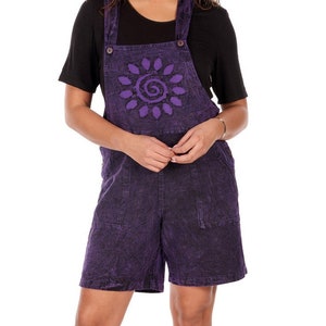 May include: A woman wearing a black t-shirt and purple overall shorts. The shorts have a sun design on the bib and pockets. She is wearing white sneakers.