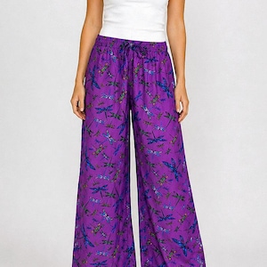 May include: A woman wearing a white tank top and wide-leg purple pants. The pants feature a dragonfly pattern in blue, green, and purple. The outfit is completed with brown sandals.