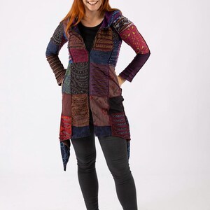 May include: A long patchwork jacket with a zip fastening. The jacket displays a variety of colours and patterns, including blue, red, purple, and brown. The sleeves are long and the jacket is worn with black trousers and white trainers.