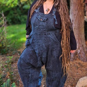 May include: Dark blue denim overalls with a bib pocket and loose, baggy legs. The overalls are worn over a black long-sleeve shirt. The overall's legs have a blue decorative design near the ankles. The person is wearing brown shoes.