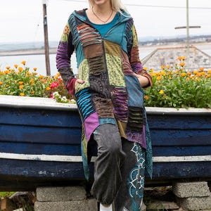May include: A colourful patchwork hooded jacket with a zip closure and pockets. The jacket features shades of blue, purple, and green. The person is wearing grey trousers and white trainers. The background includes a blue boat and flowers.
