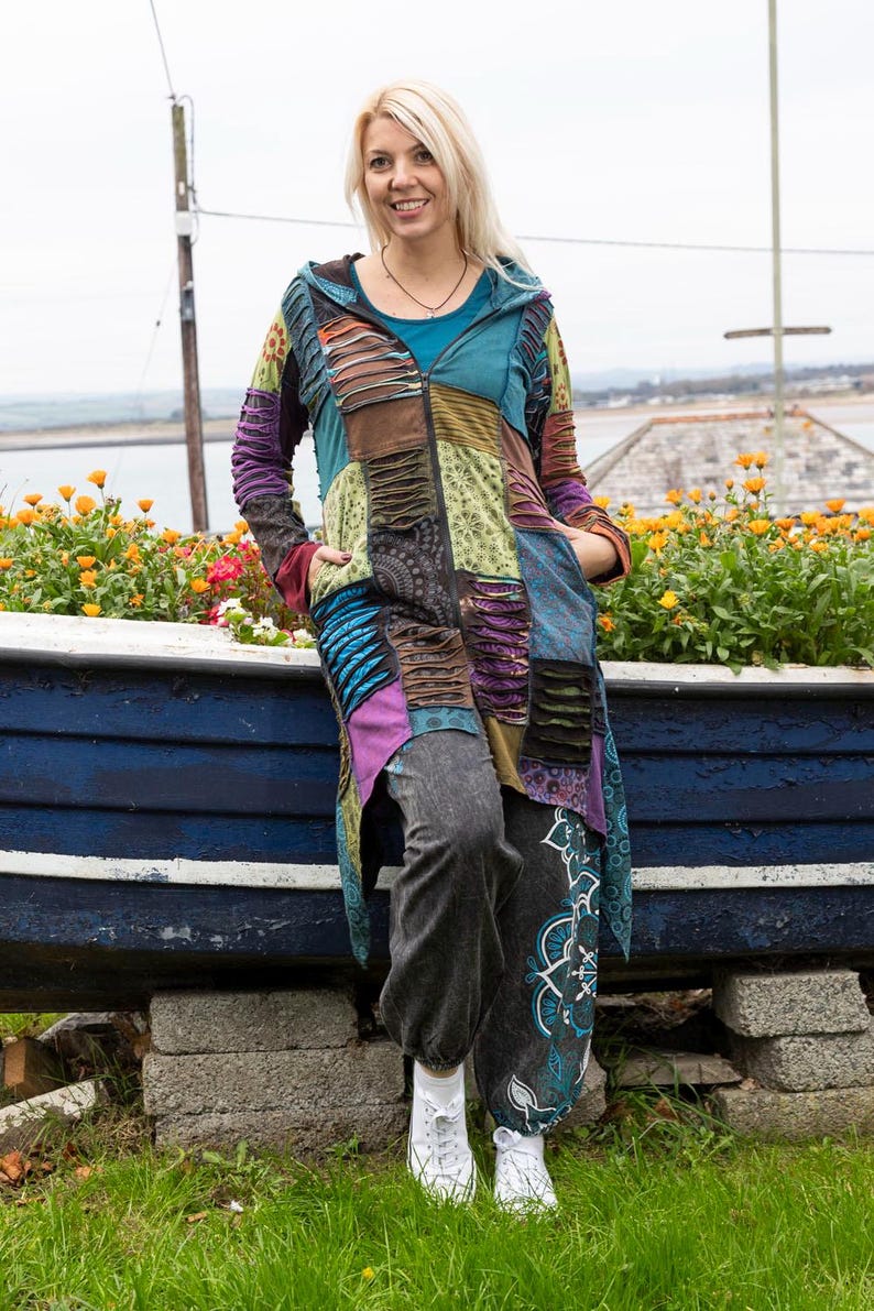 May include: A colourful patchwork hooded jacket with a zip closure and pockets. The jacket features shades of blue, purple, and green. The person is wearing grey trousers and white trainers. The background includes a blue boat and flowers.