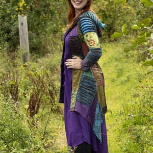 May include: A woman in a patchwork jacket and purple dress, standing in a field. The jacket has a variety of colours and patterns, including blue, green, and floral designs. The dress is a deep purple colour.