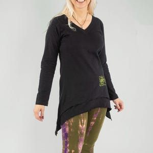 Könnte beinhalten: Schwarze Langarm-Tunika mit V-Ausschnitt und asymmetrischem Saum. Die Tunika hat ein grünes Design auf der unteren rechten Seite. Kombiniert mit olivgrünen und lila Batik-Leggings und schwarzen Stiefeln. Ein lässiges Outfit.